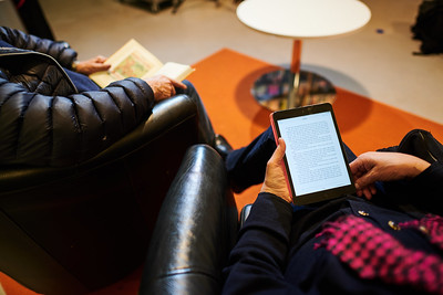 Kaksi ihmistä nojatuoleissa, näkyvät kaulasta alaspäin. Toisella on kädessä avonainen kirja, toisella e-lukulaite.