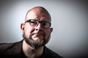 Silmälasipäinen mies katsoo kameraan. Kuva on kasvokuva. Miehellä on silmälasit ja parta.