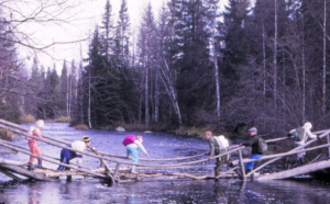 Vanha valokuva, jossa kuusi henkilöä on ylittämässä rikkonaista siltaa joen yli.